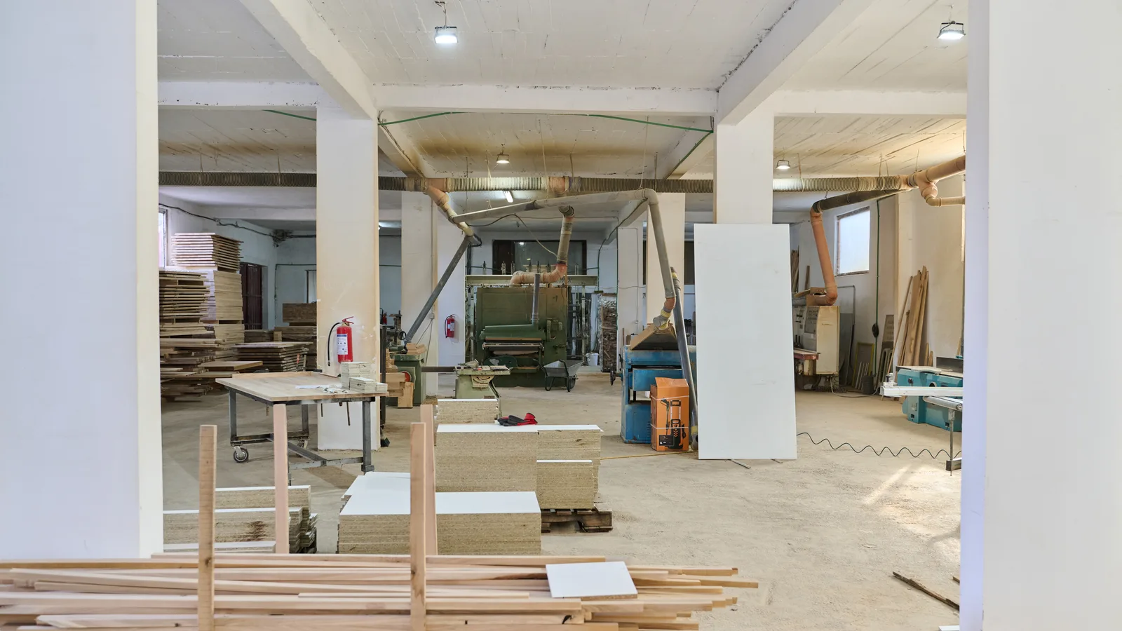 Interior of a light industrial woodworking shop with dust extraction ducts, stacked plywood and fire extinguishers mounted on the columns