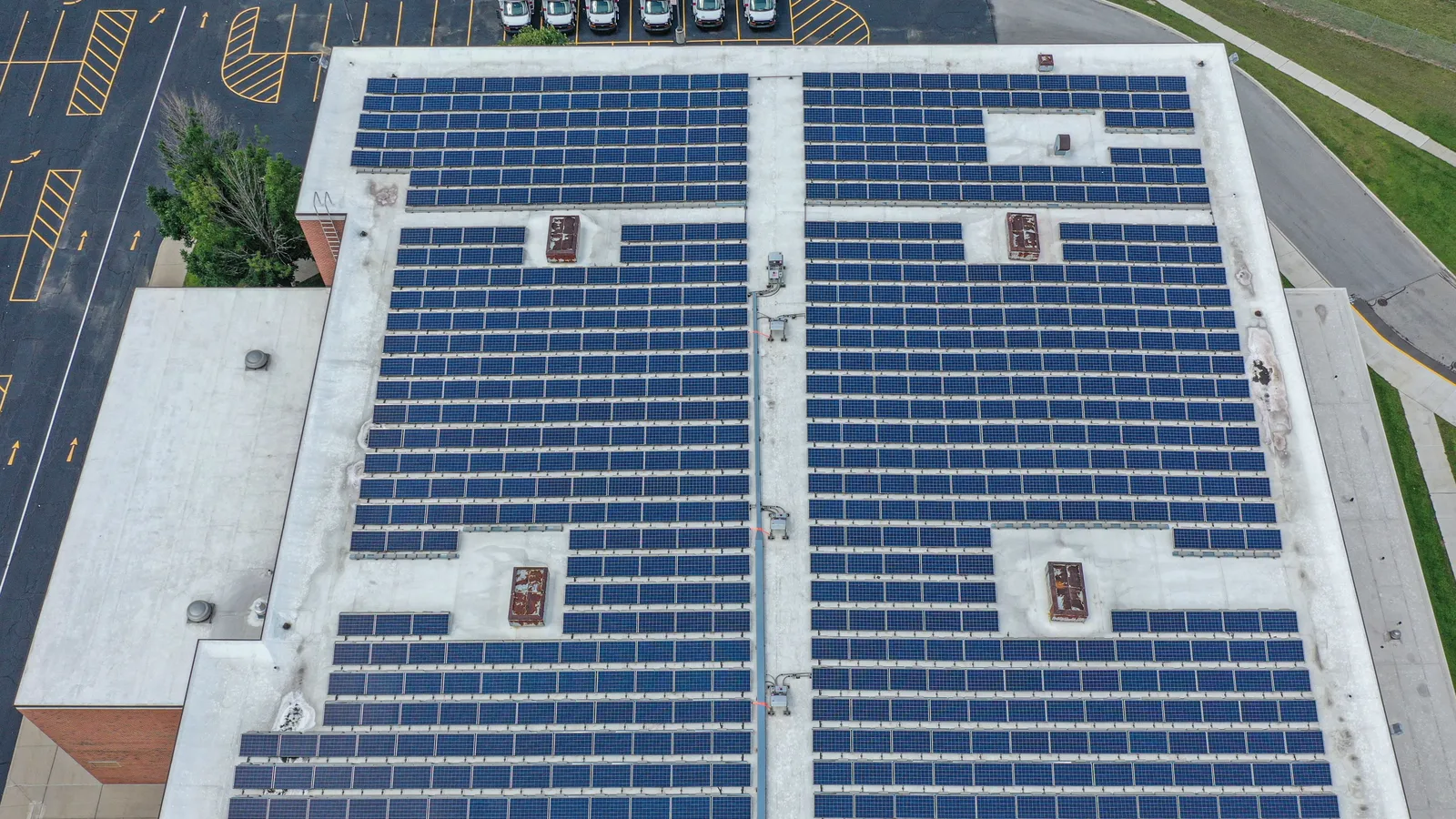 Aerial view of a flat commercial roof covered in rows of solar panels beside a parking lot with white vans