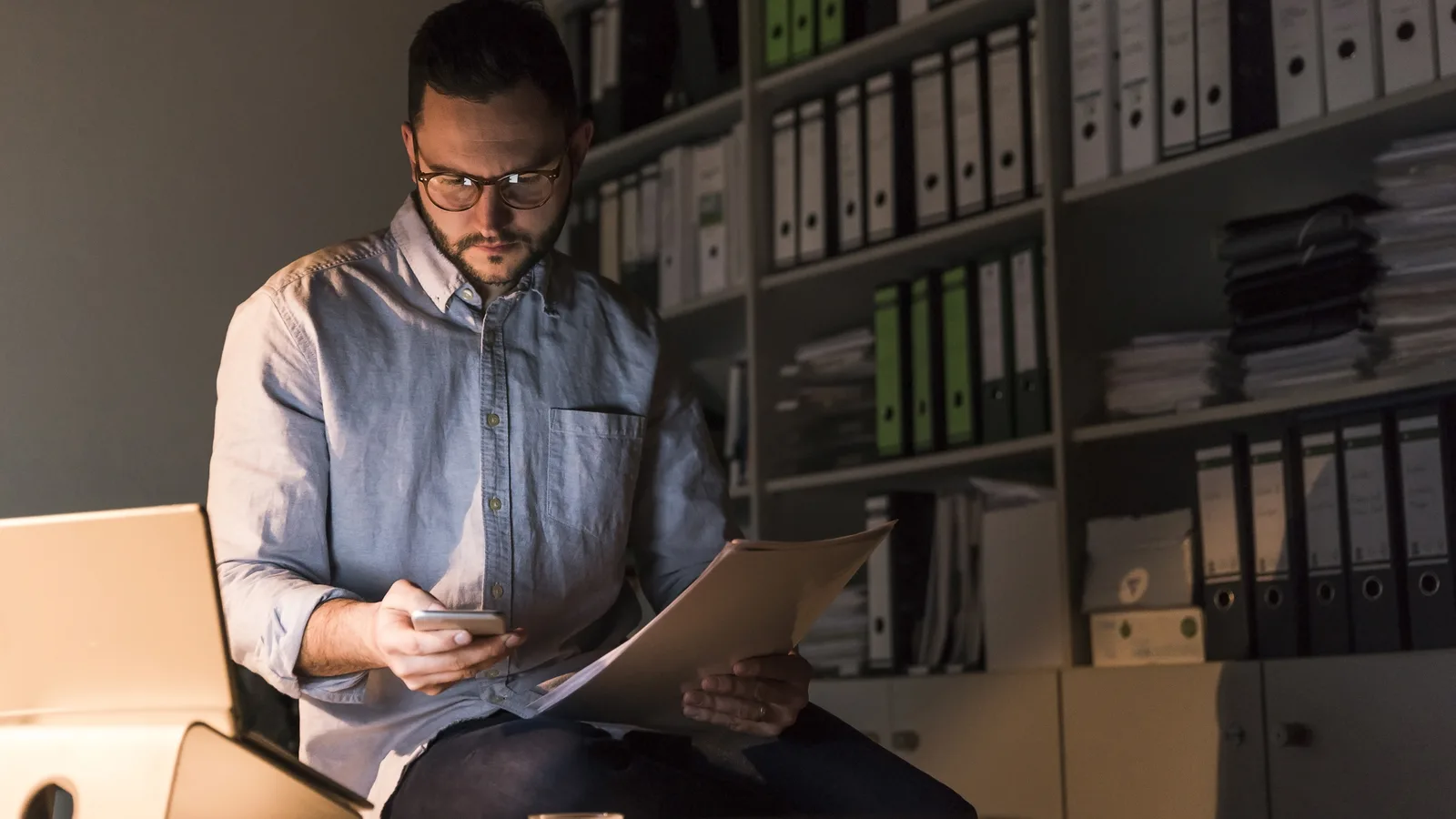 An accountant reviewing paperwork at night in an office lined with binders