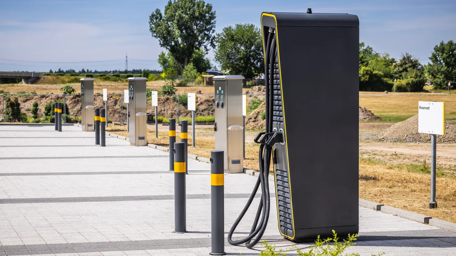 A row of electric vehicle fast chargers behind yellow and grey steel bollards on a newly paved lot