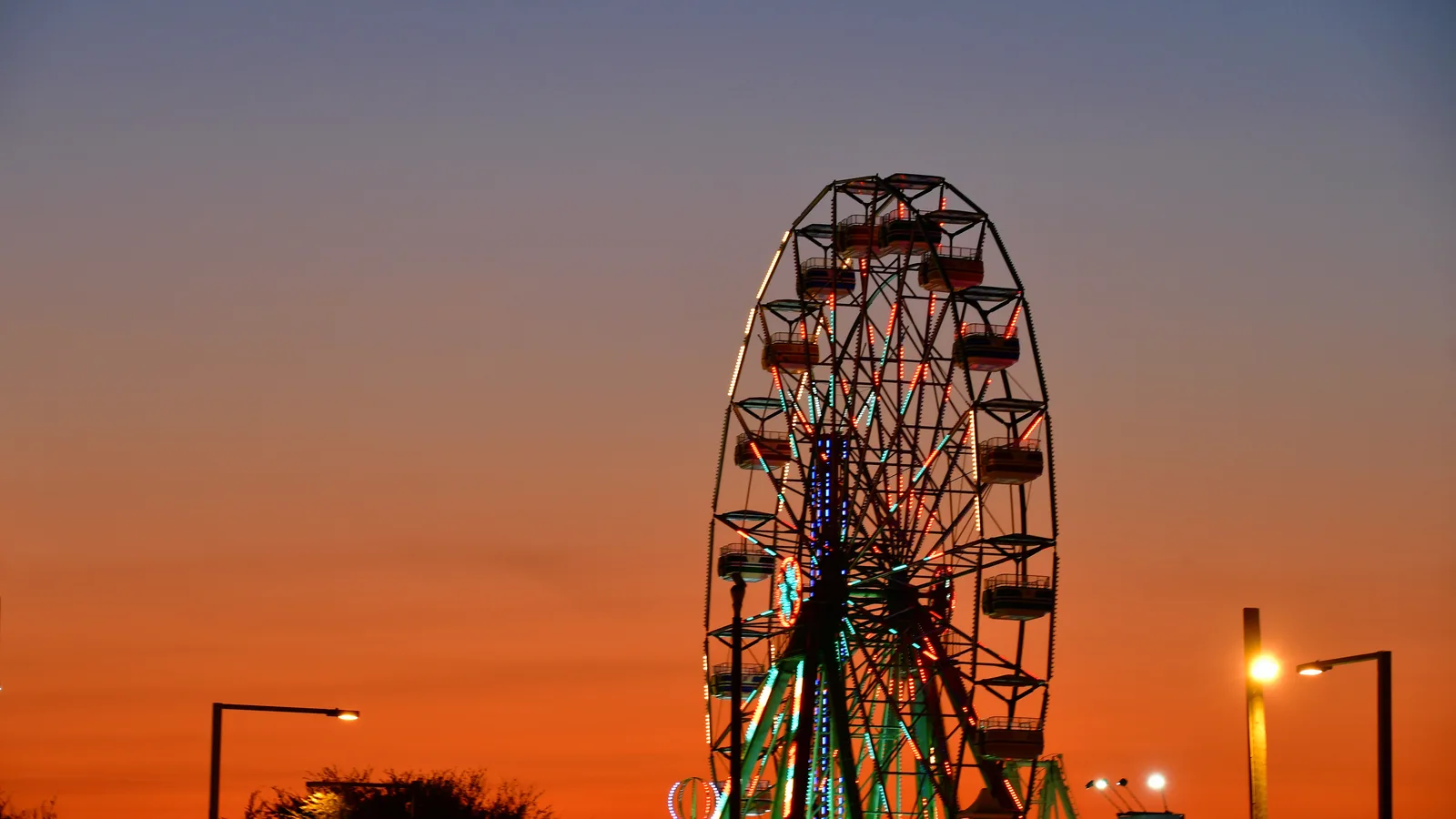 A lit ferris wheel silhouetted against an orange sunset sky at a carnival