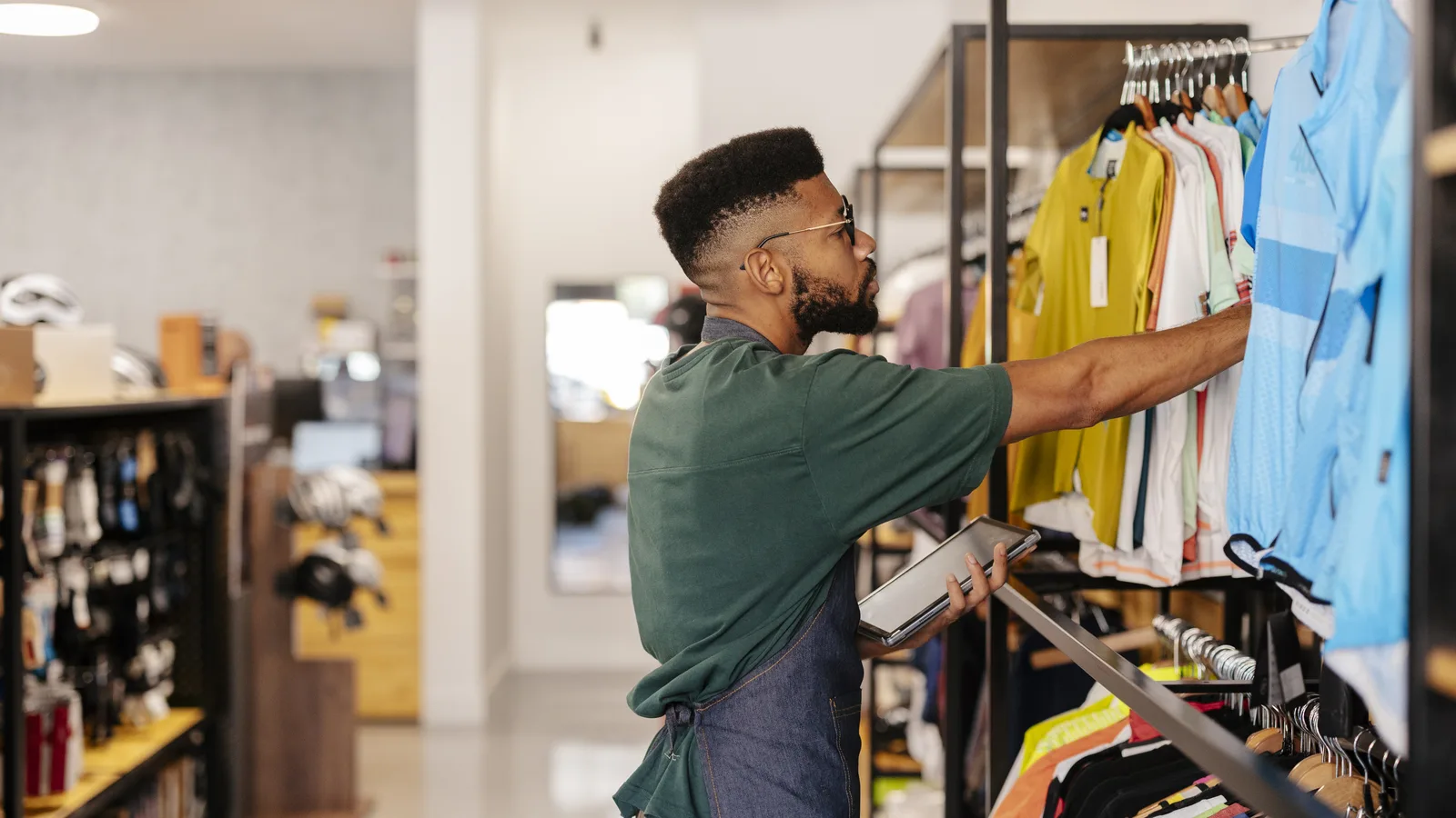 A store employee in a denim apron checks shirts on a rack while holding a tablet