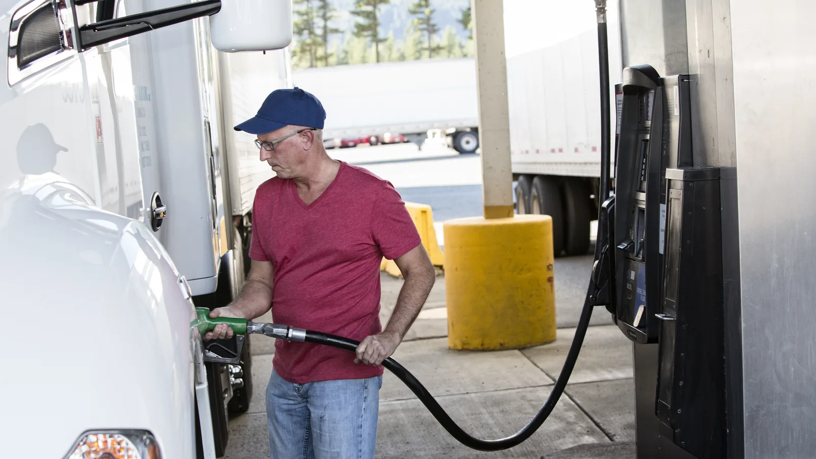 A man in a red shirt and blue cap filling a tractor trailer at a truck stop diesel pump
