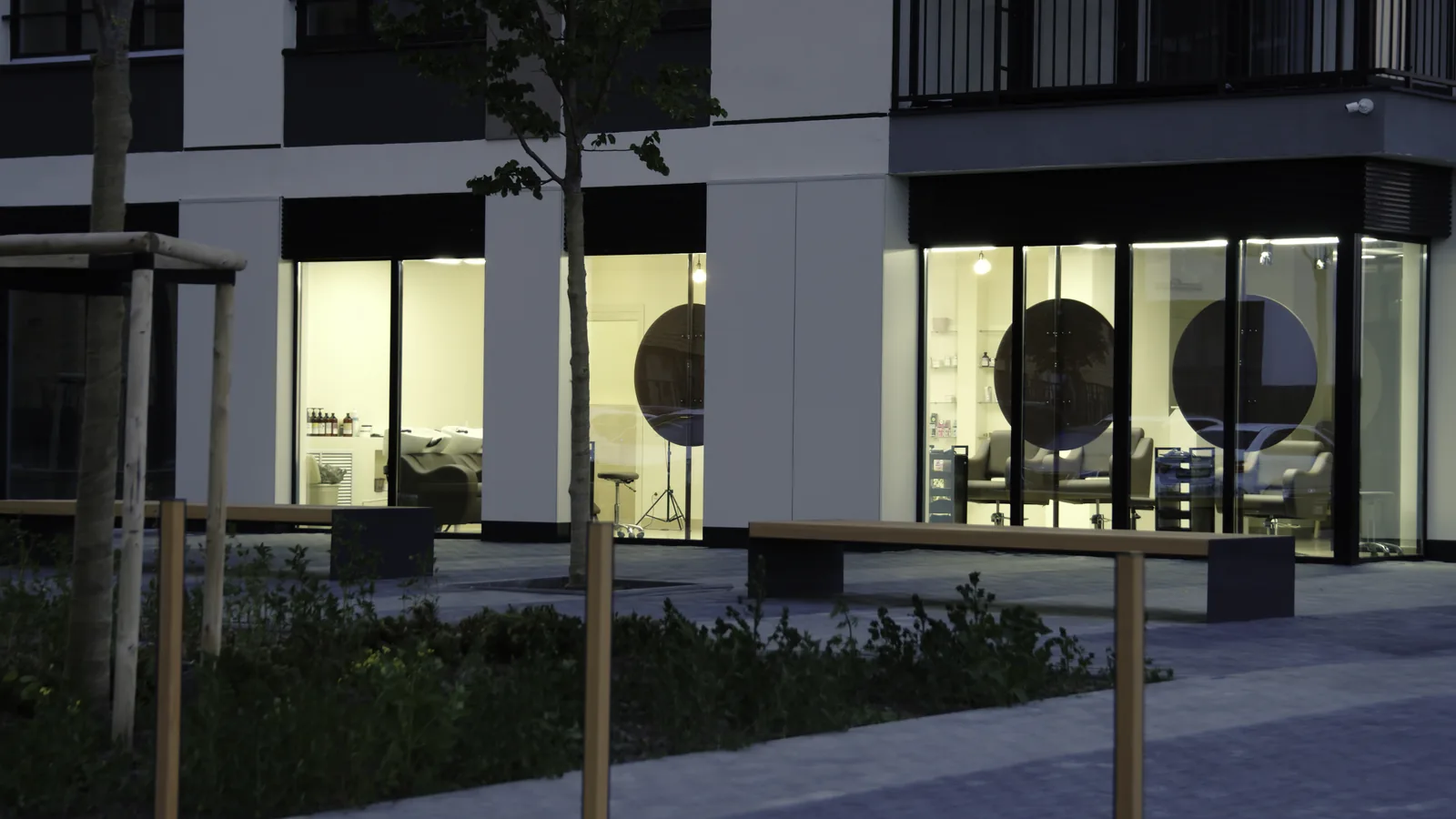Lit ground floor storefronts beneath apartments in a newly built town center at dusk