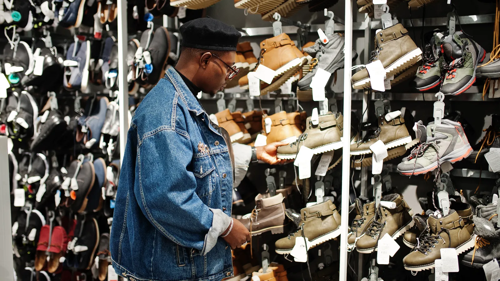 A man in a denim jacket examining boots on a densely stocked shoe store wall
