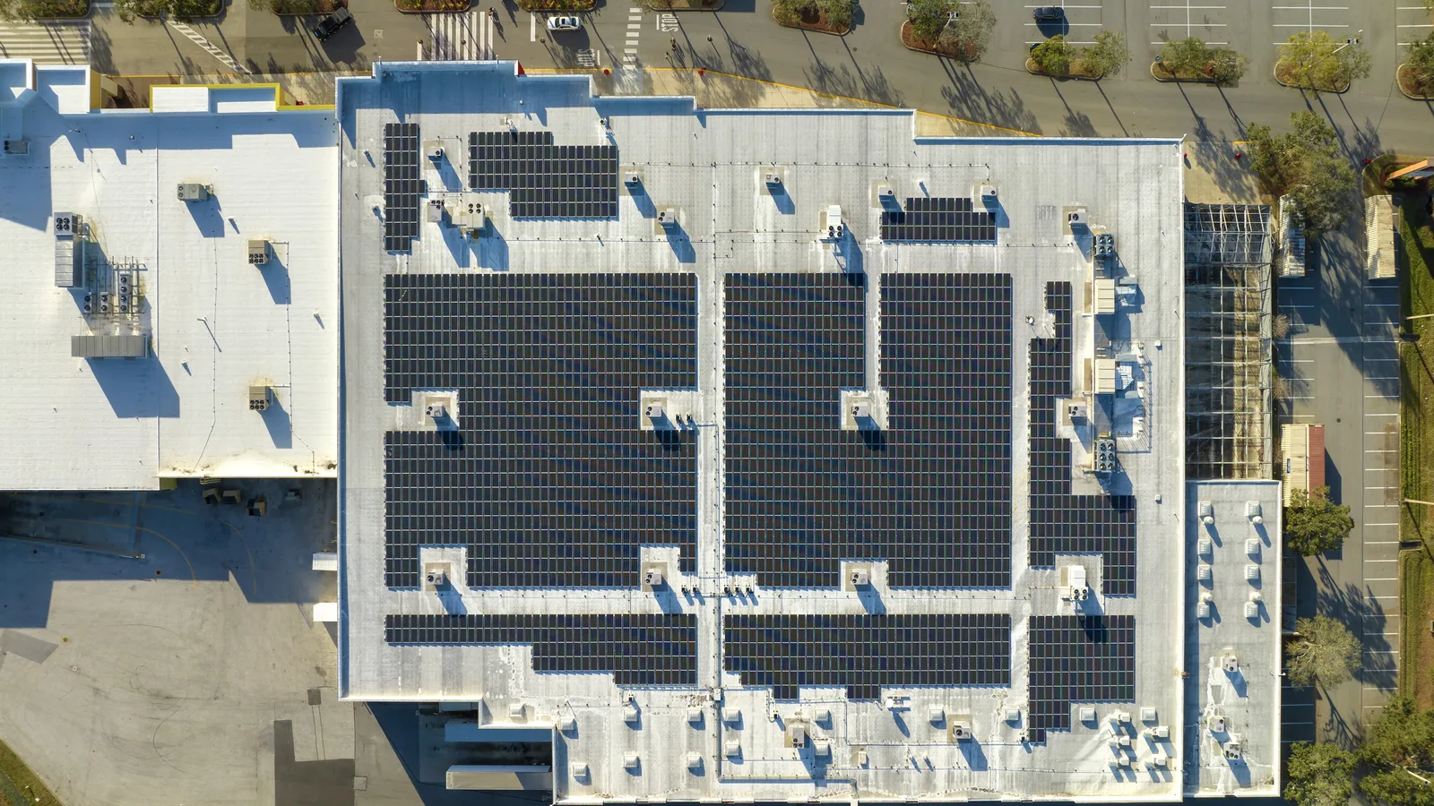 Overhead view of a flat commercial roof covered with rows of solar panels beside a parking lot