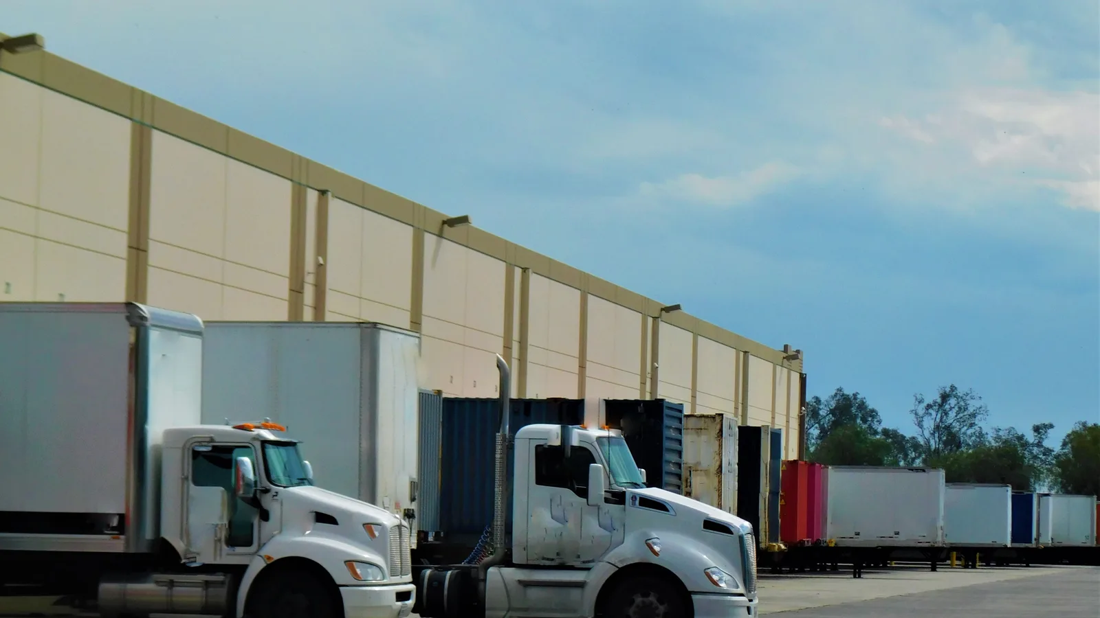 White box trucks and tractors parked at the loading docks along a long tan warehouse wall under a partly cloudy sky