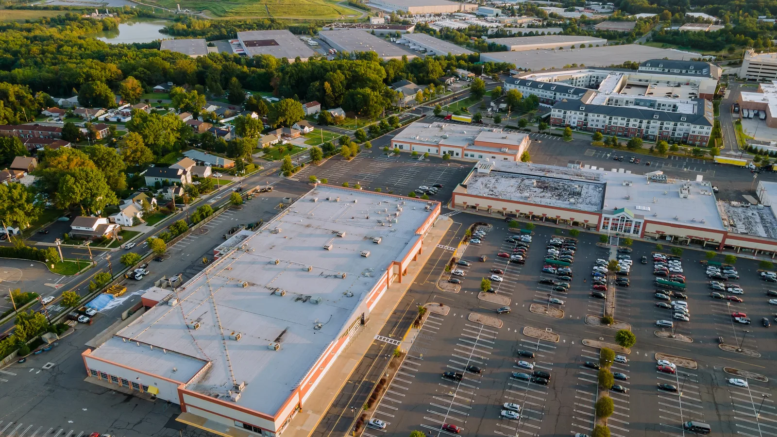 Aerial view of a suburban shopping center, its parking lot, warehouses and nearby houses in late afternoon light
